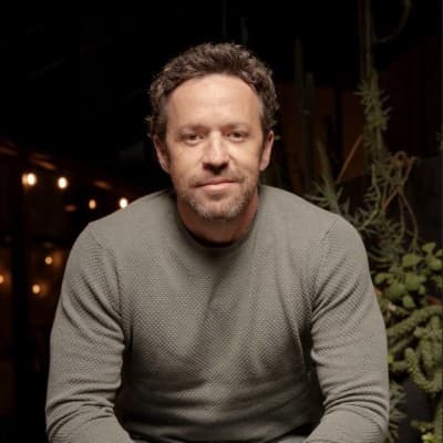 Homem de barba curta e suéter cinza texturizado em ambiente interno com iluminação suave.