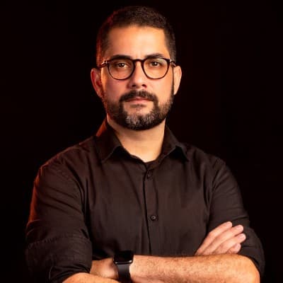 Homem barbudo com óculos e camisa preta, posando de braços cruzados em fundo preto.