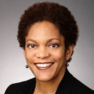 Mulher negra sorridente com cabelo afro curto, vestindo blazer preto, brincos de pérola e colar.